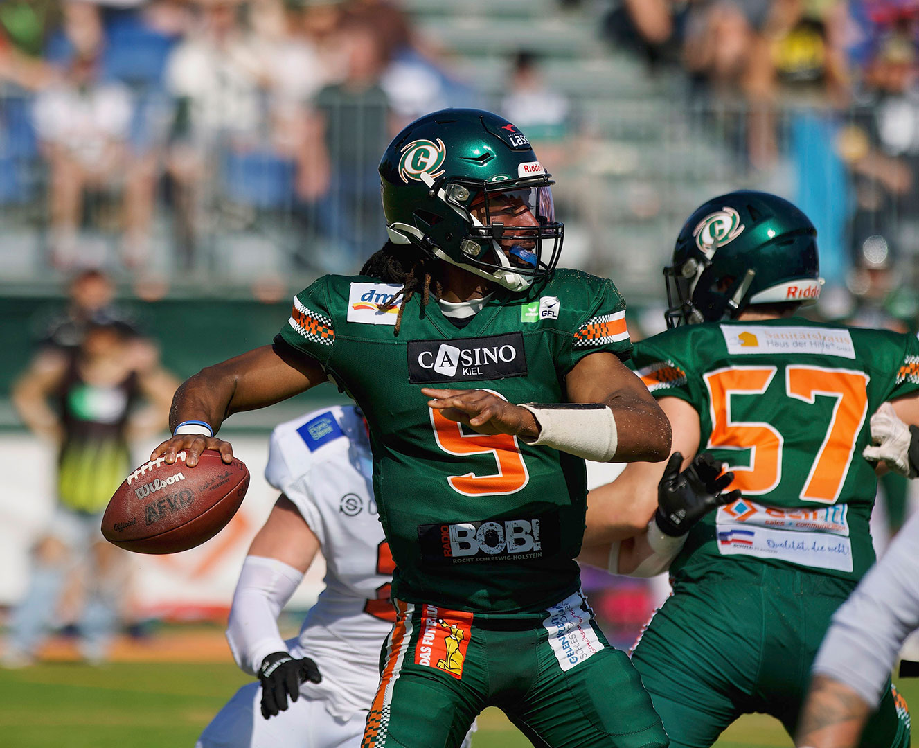 Das Foto zeigt PJ Settles, den Quarterback der Kiel Baltic Hurricanes 2025 und 2026. Auf dem Bild macht er sich gerade zum Werfen bereit und zieht den rechten Arm mit dem Football in der Hand zurück, um zum Werfen auszuholen. Er trägt die grüne Uniform der Hurricanes mit seiner orangenen Nummer 9 auf der Brust. Im Hintergrund ist Offensive Lineman Mikkel Jacobsen (Nummer 57), der einen gegnerischen Defensive Lineman von Settles abhält. Dahinter sieht man unscharf die Zuschauer auf der Tribüne im Kieler Kilia Stadion.